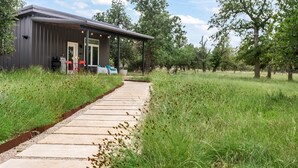 Een modern gebouw met een overdekte veranda, een pad dat er naartoe leidt, en een natuurlijk landschap met bomen en gras.