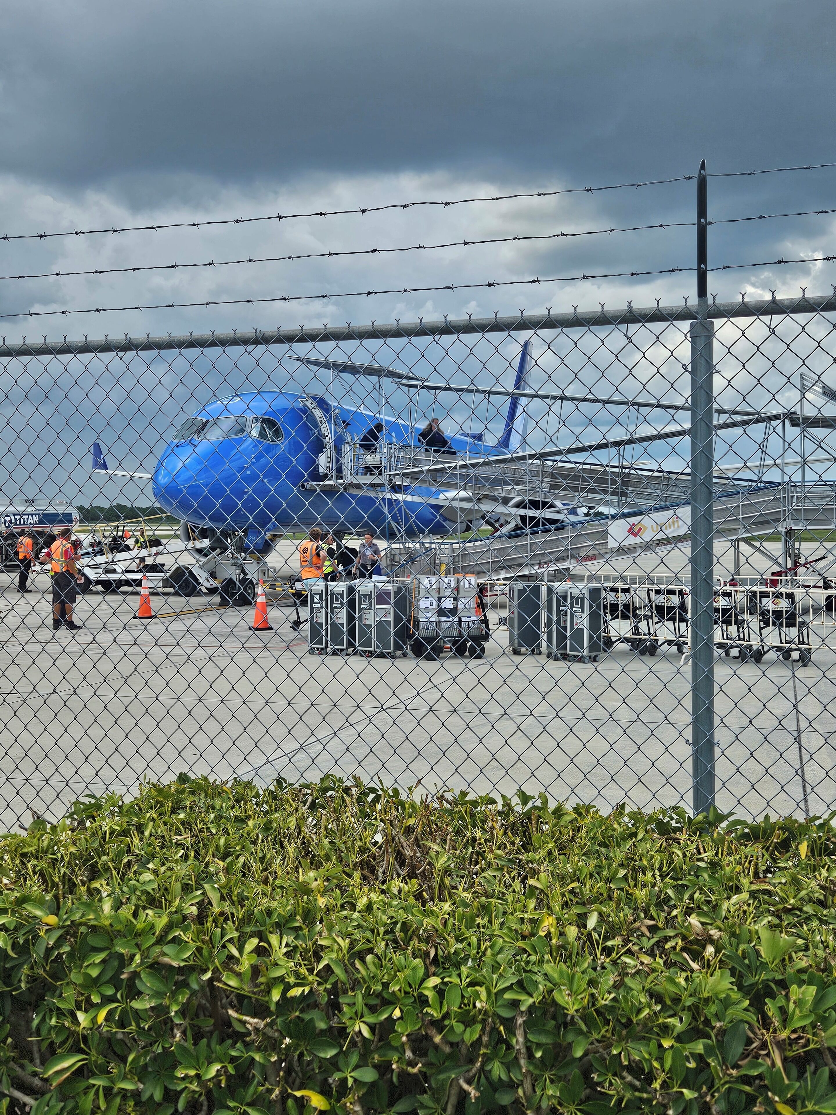 Un avion bleu est garé à une porte d’embarquement de l’aéroport, et on peut voir du personnel au sol ainsi que du matériel à proximité.