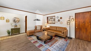A living room with a brown leather sofa, a coffee table, and a wooden floor. There is a door on the right and a wall with various decorations. - Charming 3-bedroom cabin with AC, WiFi in serene Idyllwild-Pine Cove (Idyllwild-Pine Cove)