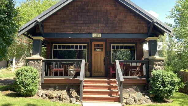 A two-story wooden house with a front porch, number 104, and a small garden area.