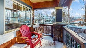 A wooden porch with a red Adirondack chair, a stone pillar, and a view of snow-covered houses.