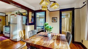 A dining area with a wooden table, chairs, and a potted plant. There is a kitchen area with a refrigerator and stove in the background.
