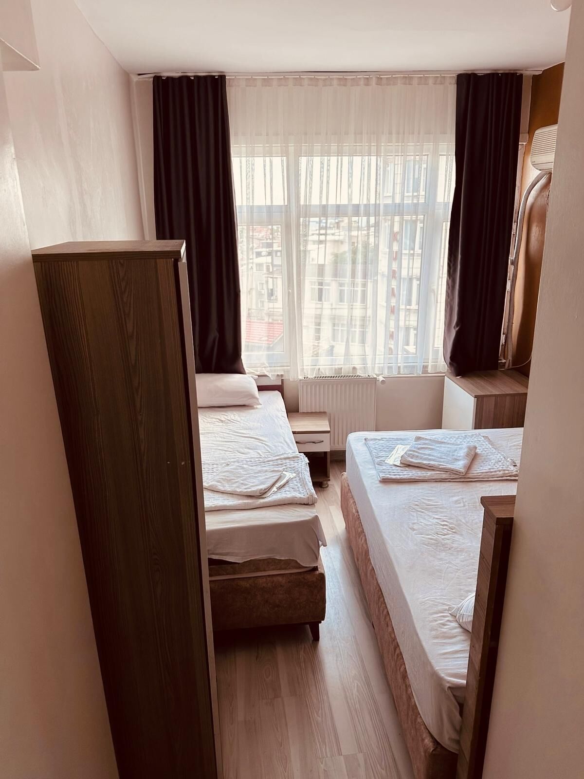 A hotel room with two beds, a wooden wardrobe, a radiator, and a window with curtains.