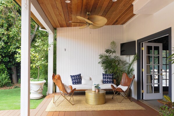 A covered outdoor seating area with a ceiling fan, a sofa with patterned cushions, two leather chairs, and a round coffee table.