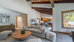 A spacious living room with a large sofa, a wooden coffee table, and a view of the kitchen and outdoor area through a large window. - Carnelian Crest, modern 3BR cabin with hot tub - perfect Couples Retreat! (Carnelian Bay)