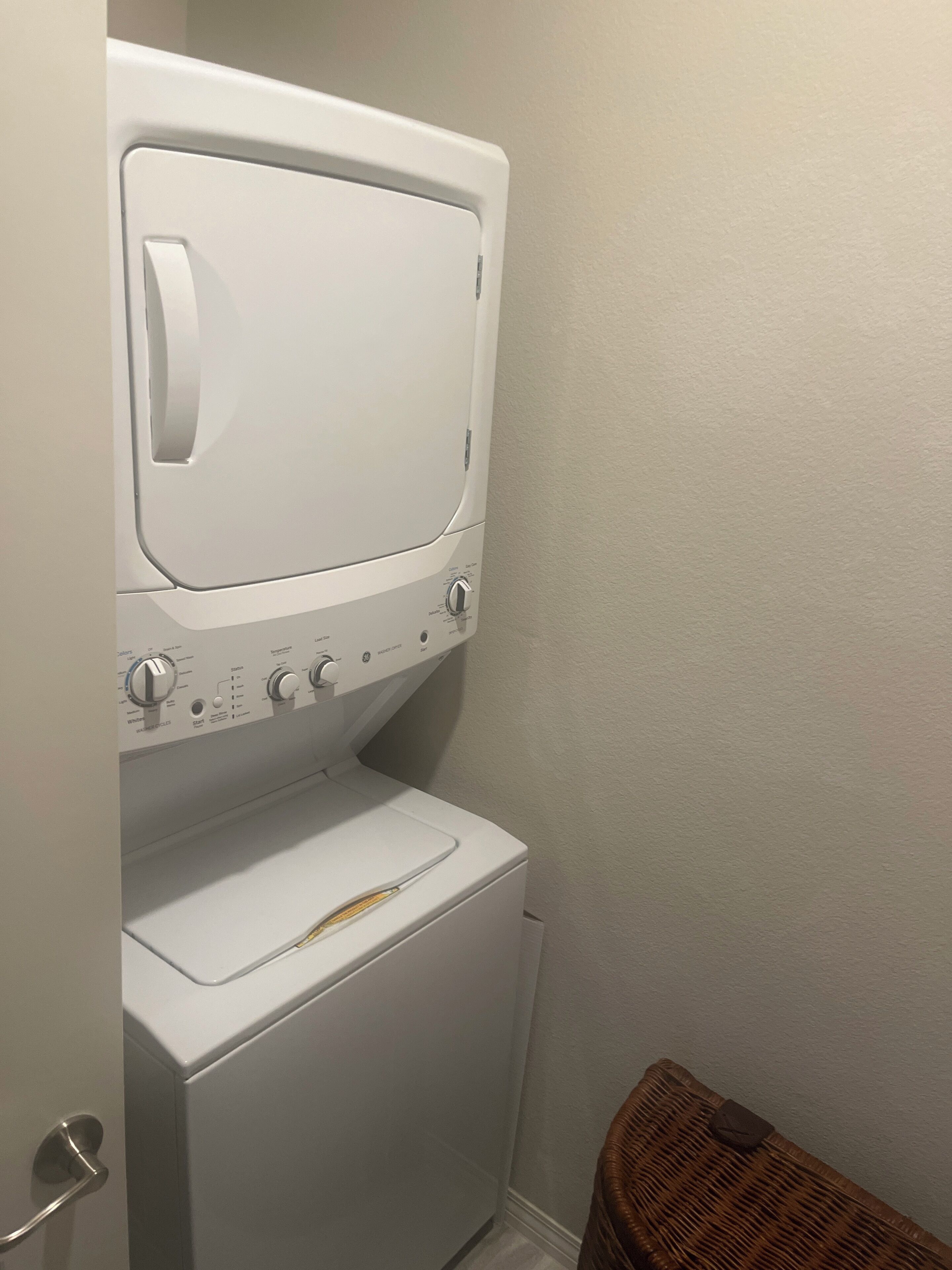 A compact laundry room with a front-loading washer and dryer, a door handle, and a wicker basket.