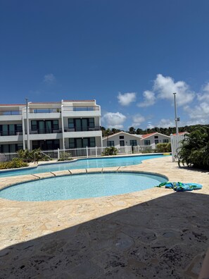 A swimming pool area with a white fence, a building with balconies, and a clear blue sky.