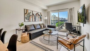 Un salon moderne avec un canapé, une table basse et des fauteuils, un balcon offrant une vue sur les immeubles et des œuvres d’art accrochées aux murs.