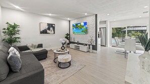 A modern living room with a grey sofa, a coffee table, and a TV. There is a balcony with a pool and outdoor seating. - New Home w/ Big Patio / Heated Pool / Pool table / Ping pong table (Miami Gardens)