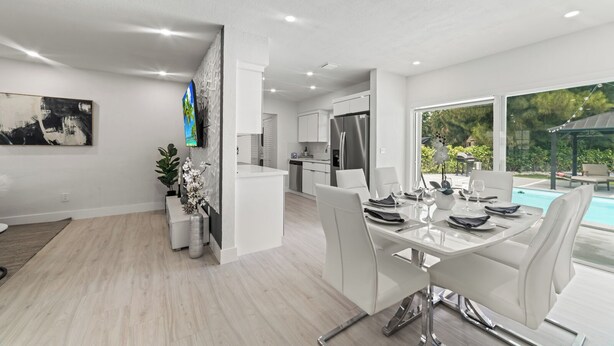 A modern kitchen with white cabinets and stainless steel appliances, a dining area with white chairs and a glass table, and a view of a swimming pool. - New Home w/ Big Patio / Heated Pool / Pool table / Ping pong table (Miami Gardens)