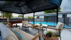 A covered outdoor seating area with a pool in the background. - New Home w/ Big Patio / Heated Pool / Pool table / Ping pong table (Miami Gardens)