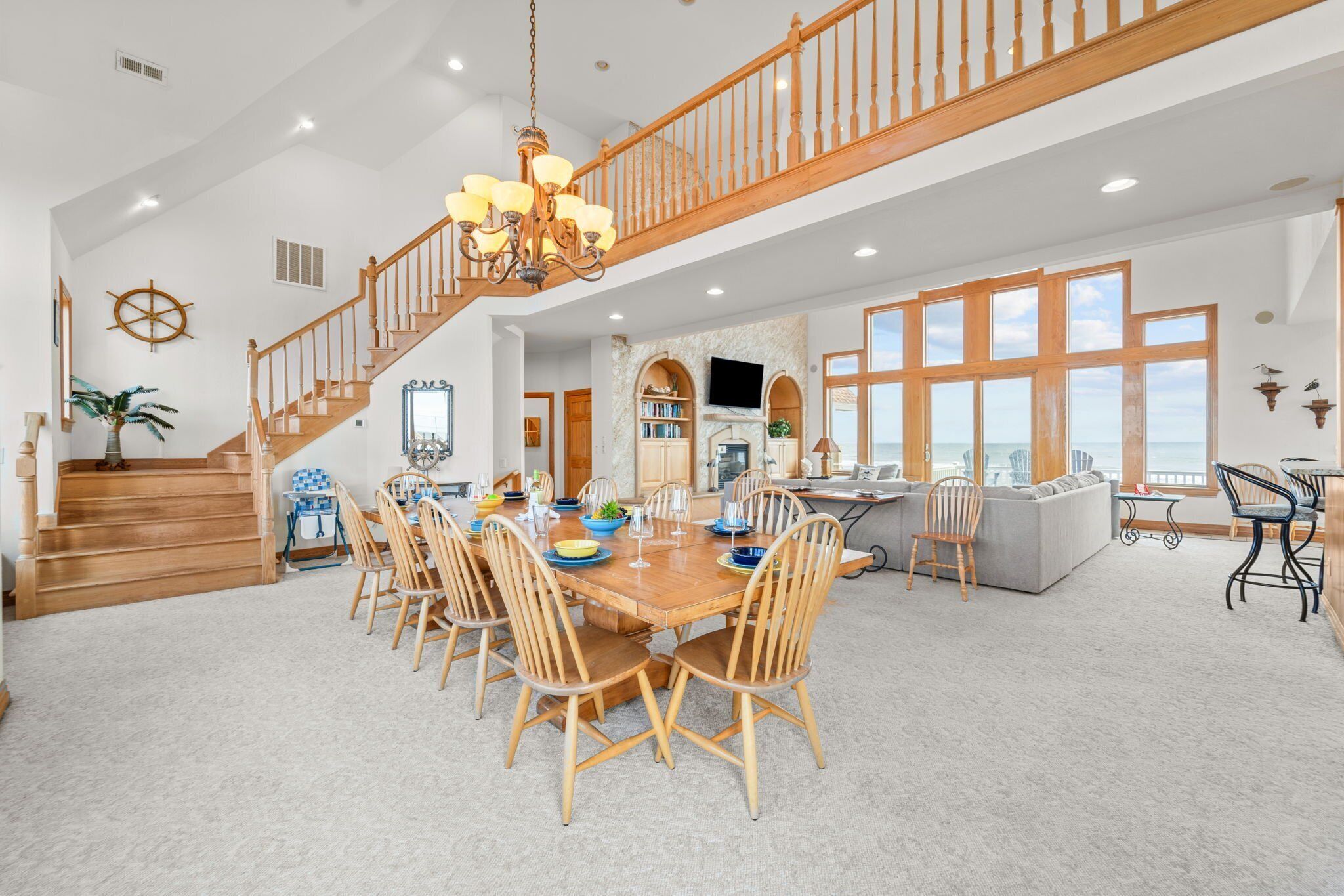 A spacious dining area with a wooden table and chairs, a staircase with wooden railings, and a view of the ocean through large windows.