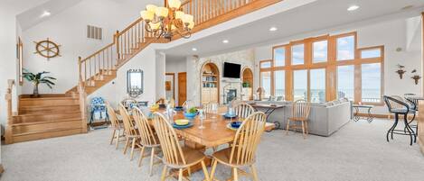A spacious dining area with a wooden table and chairs, a staircase with wooden railings, and a view of the ocean through large windows.