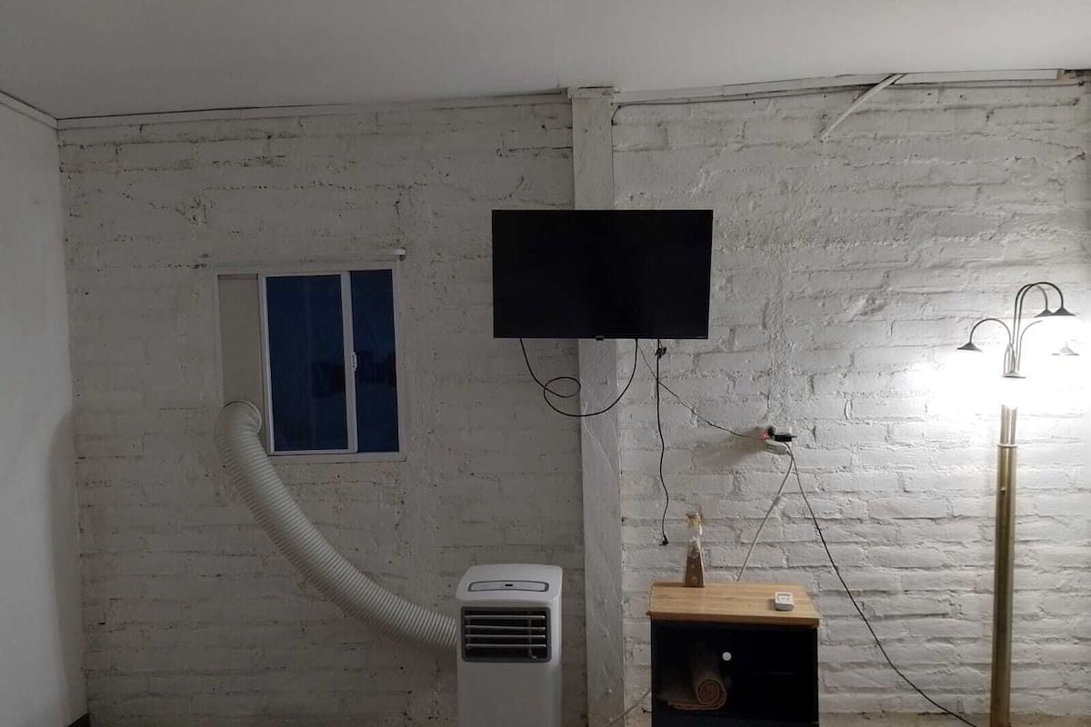 Una habitación con pared de ladrillo blanco, una ventana pequeña, un televisor montado, un calentador portátil y una lámpara de pie.