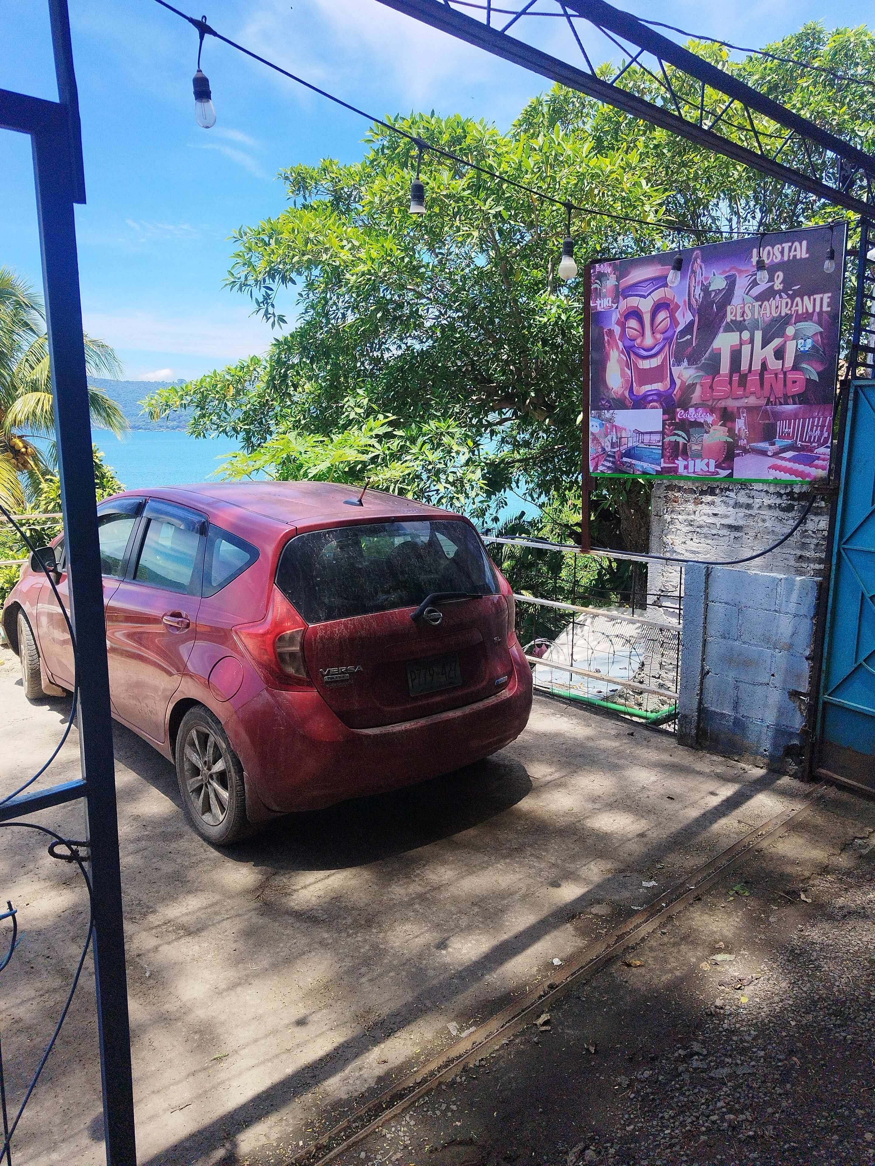 Un coche rojo estacionado en una zona sombreada, con un cartel que anuncia un restaurante llamado 'Tiki Island'.