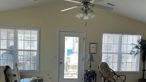 Ein geräumiges Wohnzimmer mit Deckenventilator, einem Schaukelstuhl, einem Sessel mit Beistelltisch und Blick durch eine Tür nach draußen.