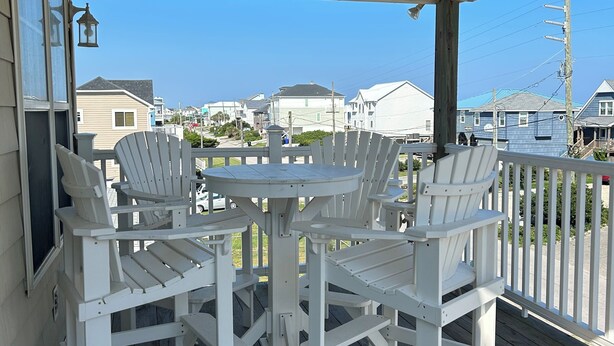 Een overdekt terras met witte Adirondack-stoelen en een ronde tafel, met uitzicht op een woonwijk.