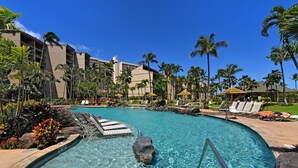 A pool area with clear water, lounge chairs, palm trees, and a building in the background.