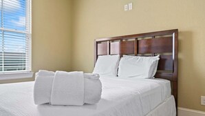 Una cama bien tendida con sábanas y toallas blancas, un cabecero de madera y una ventana con persianas.