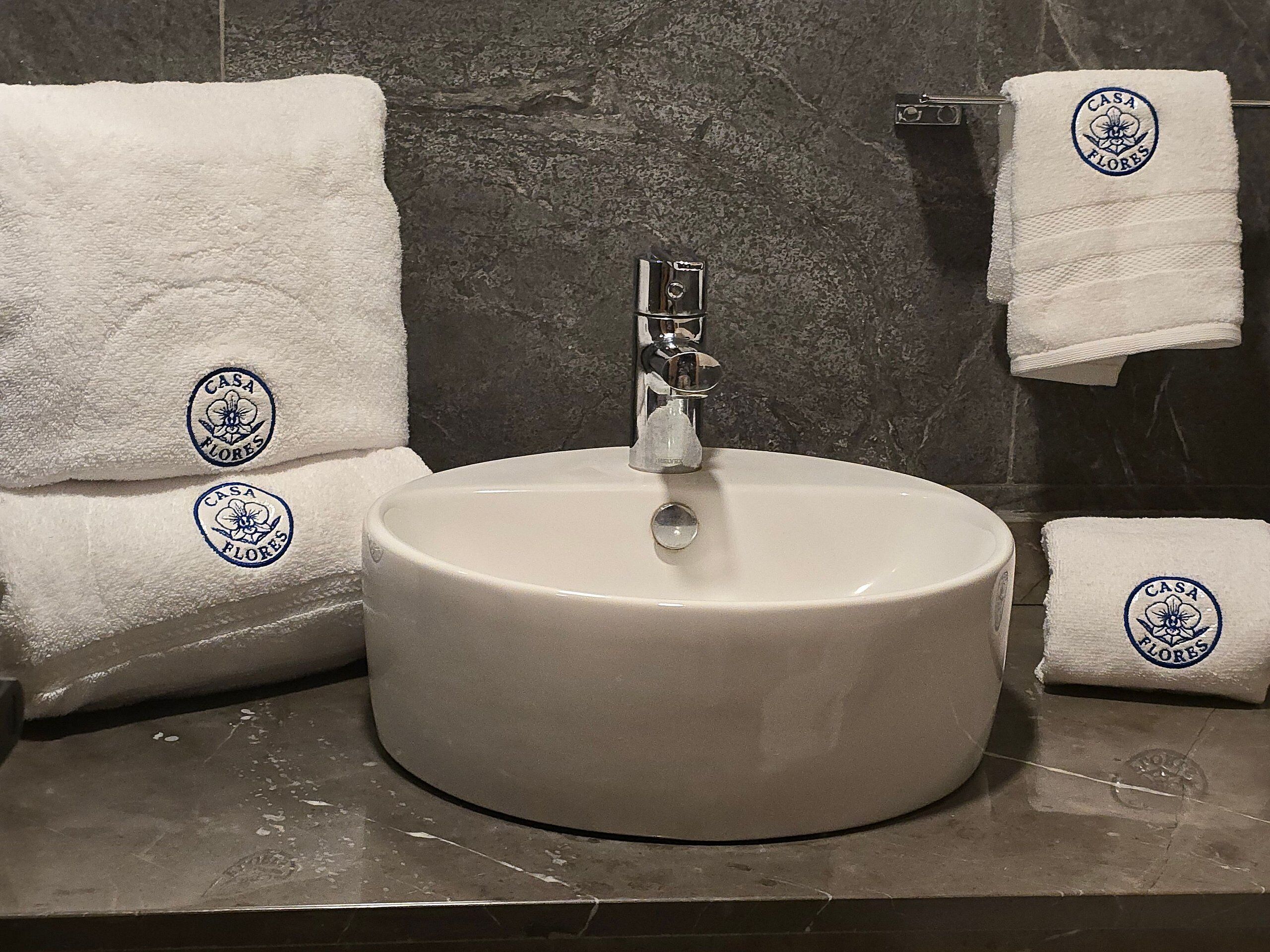A bathroom with a round white sink, a mirror, and two towels with a logo.
