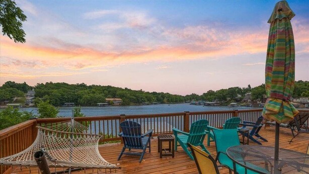 A wooden deck with outdoor furniture, including chairs and a table, overlooking a body of water with a view of a forested area in the distance. - Lakefront Fishing Retreat w/ Dock, Hot Tub & Deck Views (Sunrise Beach)