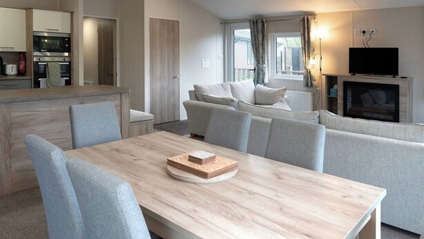 A modern living room with a wooden dining table, grey upholstered sofas, a flat-screen TV, and a fireplace. - Bittern Lodge: 3BD Retreat in South Lakeland Park (Borwick)