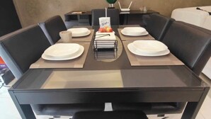 Un coin repas avec une table rectangulaire dressée pour quatre personnes, entourée de chaises noires. Une horloge murale est visible au-dessus de la table.