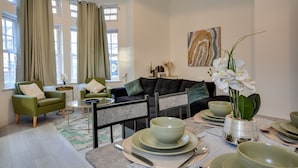 A modern living room with a sofa, chairs, a dining table set with plates and bowls, and a vase with flowers. - Cosy Wigan Apartment With Sunny Views (Wigan)