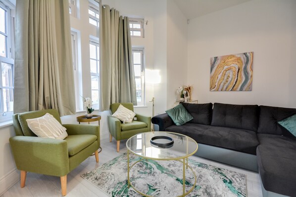 A living room with a green armchair, a black sofa, a glass coffee table, and a marble-patterned rug.