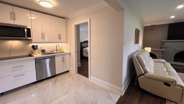 A modern kitchen with white cabinets, a built-in microwave, and a dishwasher. There is a living area with a beige sofa and a fireplace.