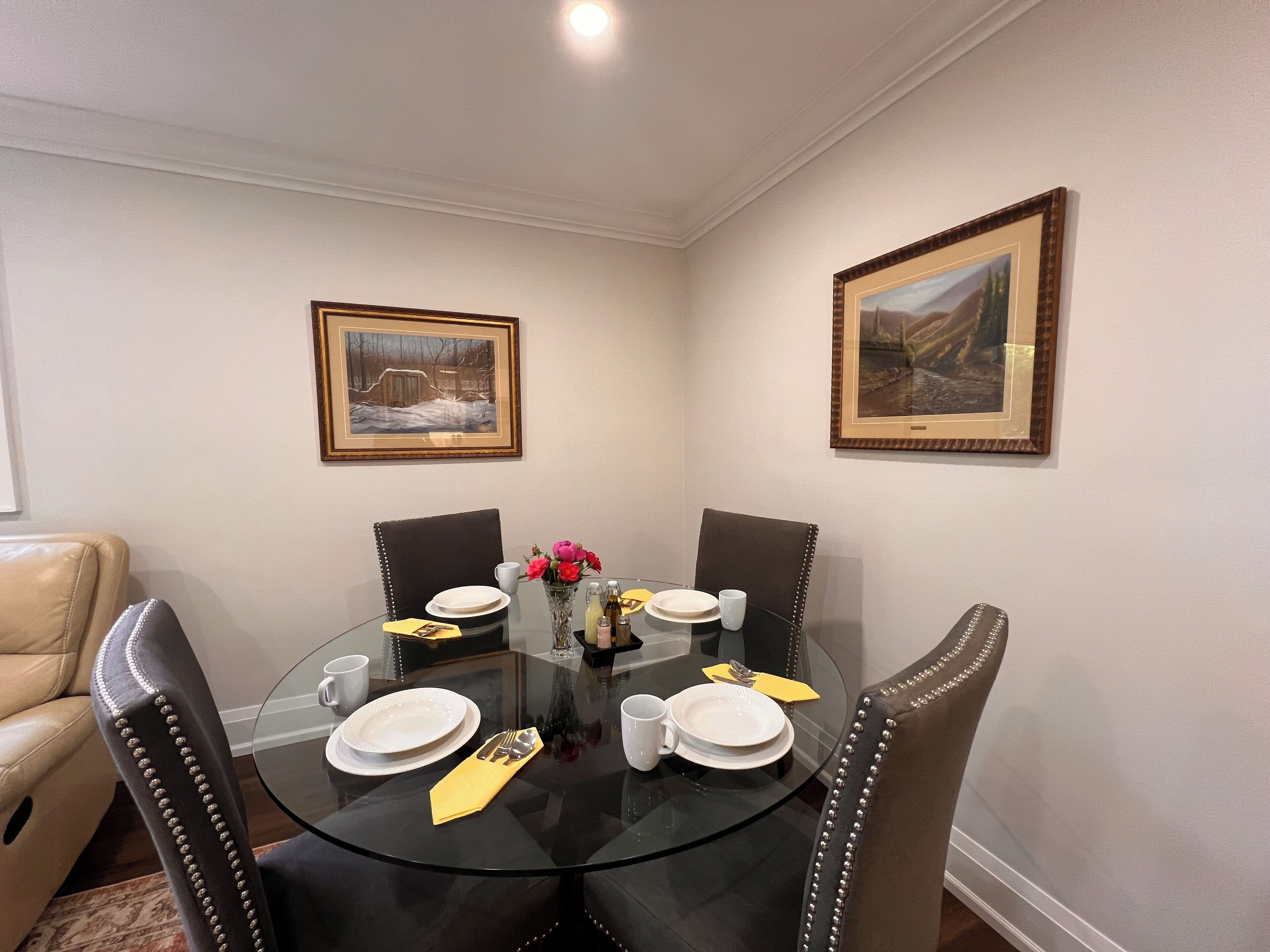 A dining area with a round glass table, four chairs, and two framed pictures on the walls.