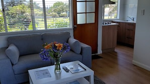 Una sala moderna con un sofá gris, una mesa de centro blanca con un florero, y vista a la cocina a través de una puerta.