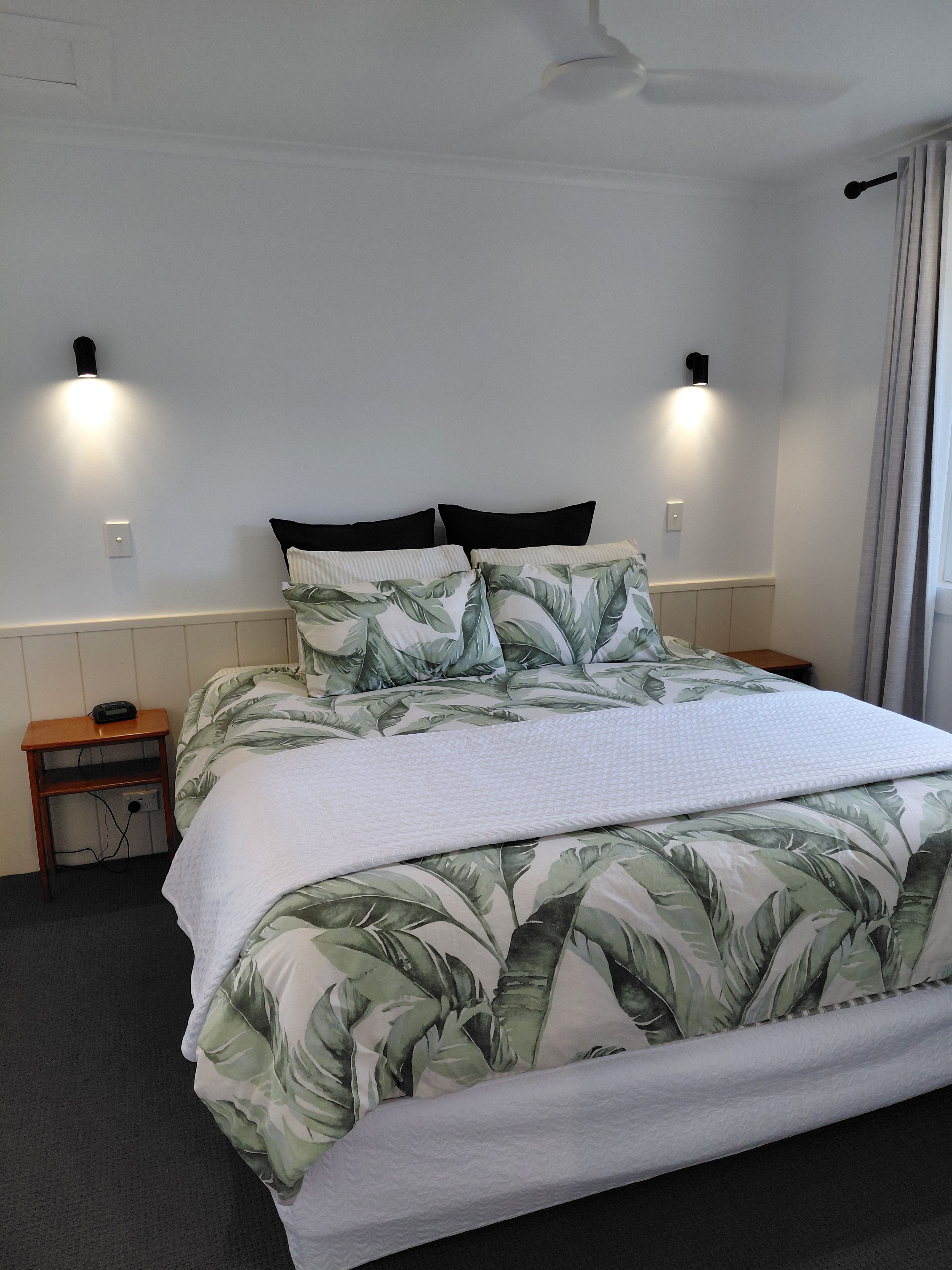 A bed with a leaf-patterned bedspread, two bedside lamps, a nightstand with a phone, and a ceiling fan.