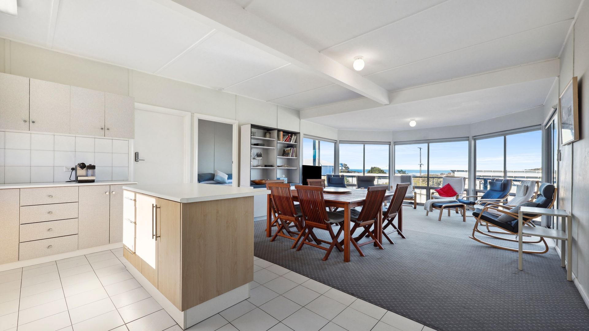 A modern kitchen with white cabinets, a dining area with wooden chairs, and a large window overlooking the sea.