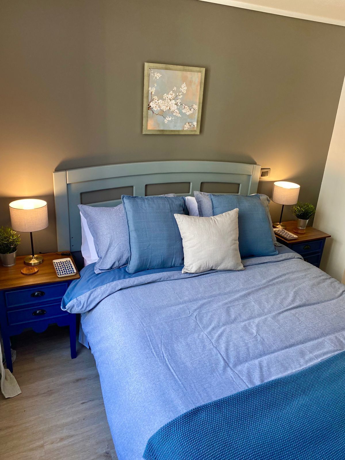 Een net opgemaakt bed met een blauw-wit kleurenschema, met aan weerszijden nachtkastjes met lampen en een kleine plant.