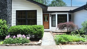 Uma casa com fachada de pedra, varanda frontal e um jardim bem cuidado com diversas plantas e um caminho que leva à porta.