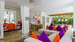 A spacious living area with a sofa set, a glass coffee table, and a kitchen in the background.