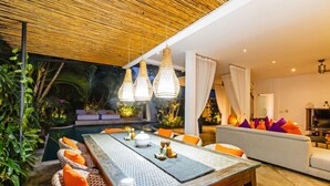 A modern outdoor dining area with a glass table, wicker chairs, and orange cushions. There is a pool in the background, and the space is surrounded by lush greenery.