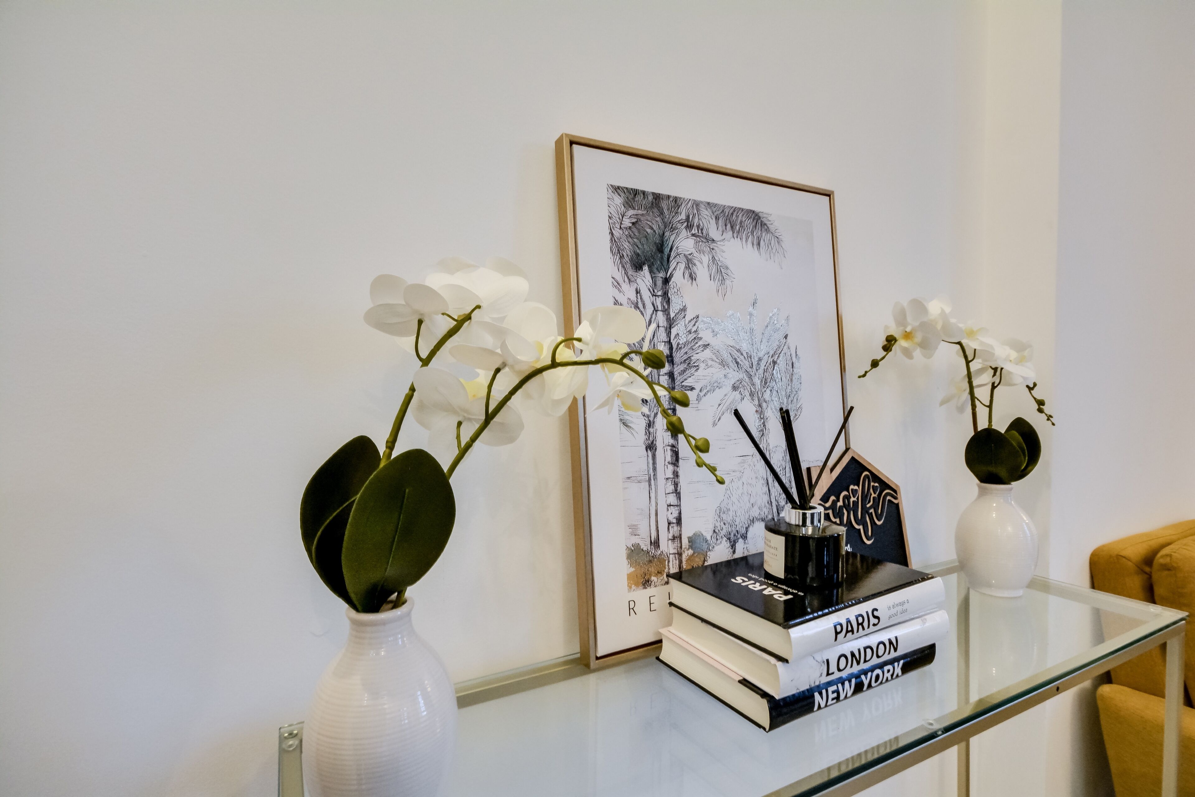 A glass table with books titled 'Paris,' 'London,' and 'New York,' a framed black and white photograph, and two white vases with orchids.