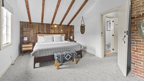 A bedroom with a wooden ceiling, a bed with a checkered bedspread, a bedside table with a lamp, and a small bench.