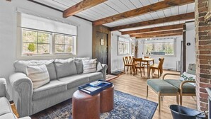A spacious living area with a grey sofa, wooden dining table, and chairs. There are large windows providing natural light and a view of the outdoors.