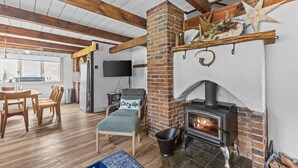 A cozy living room with a brick fireplace, wooden beams, a comfortable lounge chair, and a dining area.