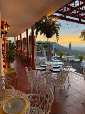 An outdoor dining area with white ornate chairs and tables, a pergola with hanging lights, and a view of the mountains at sunset. - HOTEL BALCON DEL CIELO  (Dosquebradas)