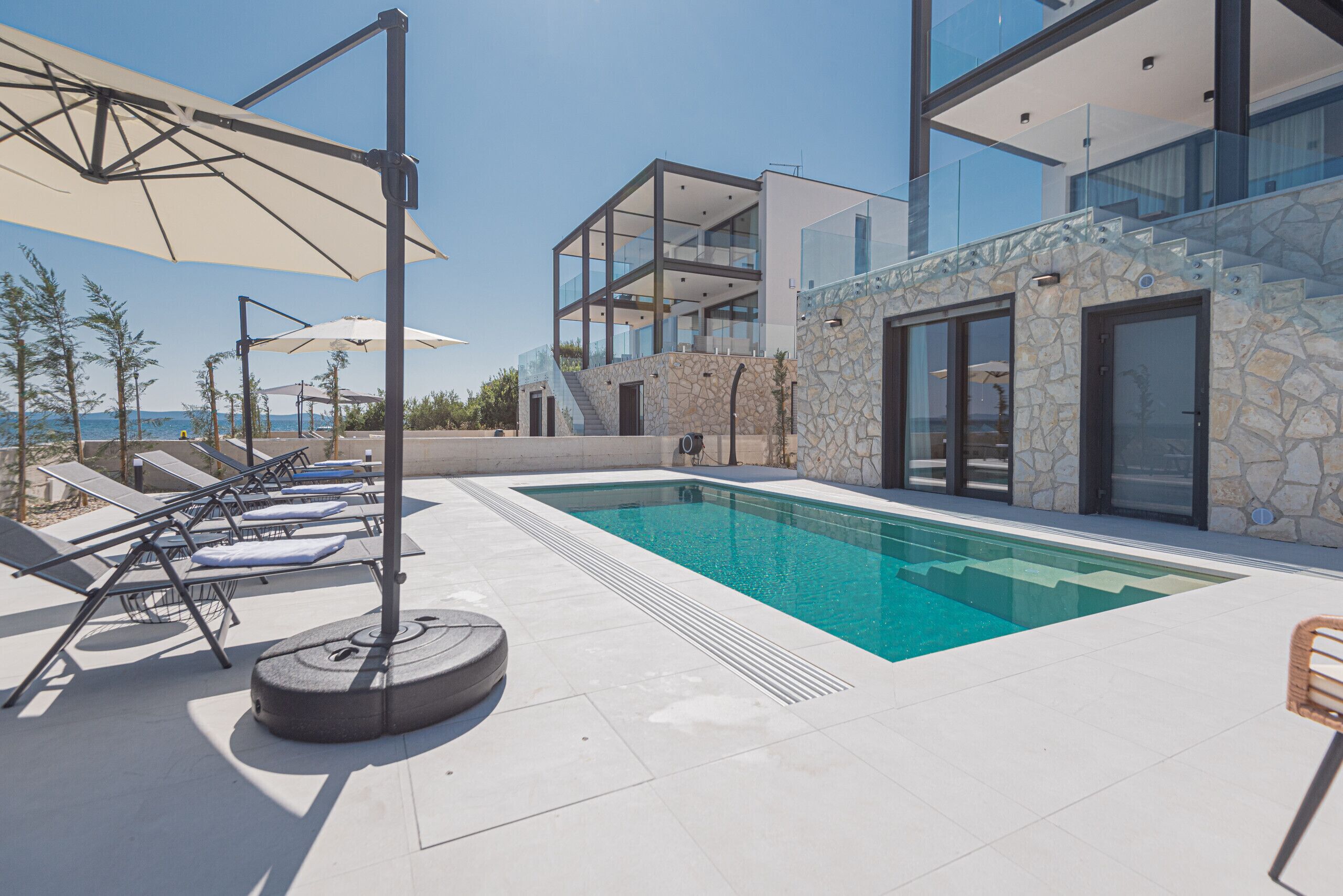 Un espace piscine extérieur moderne, doté de chaises longues et de parasols, d’un mur en pierre et d’un bâtiment entièrement vitré.