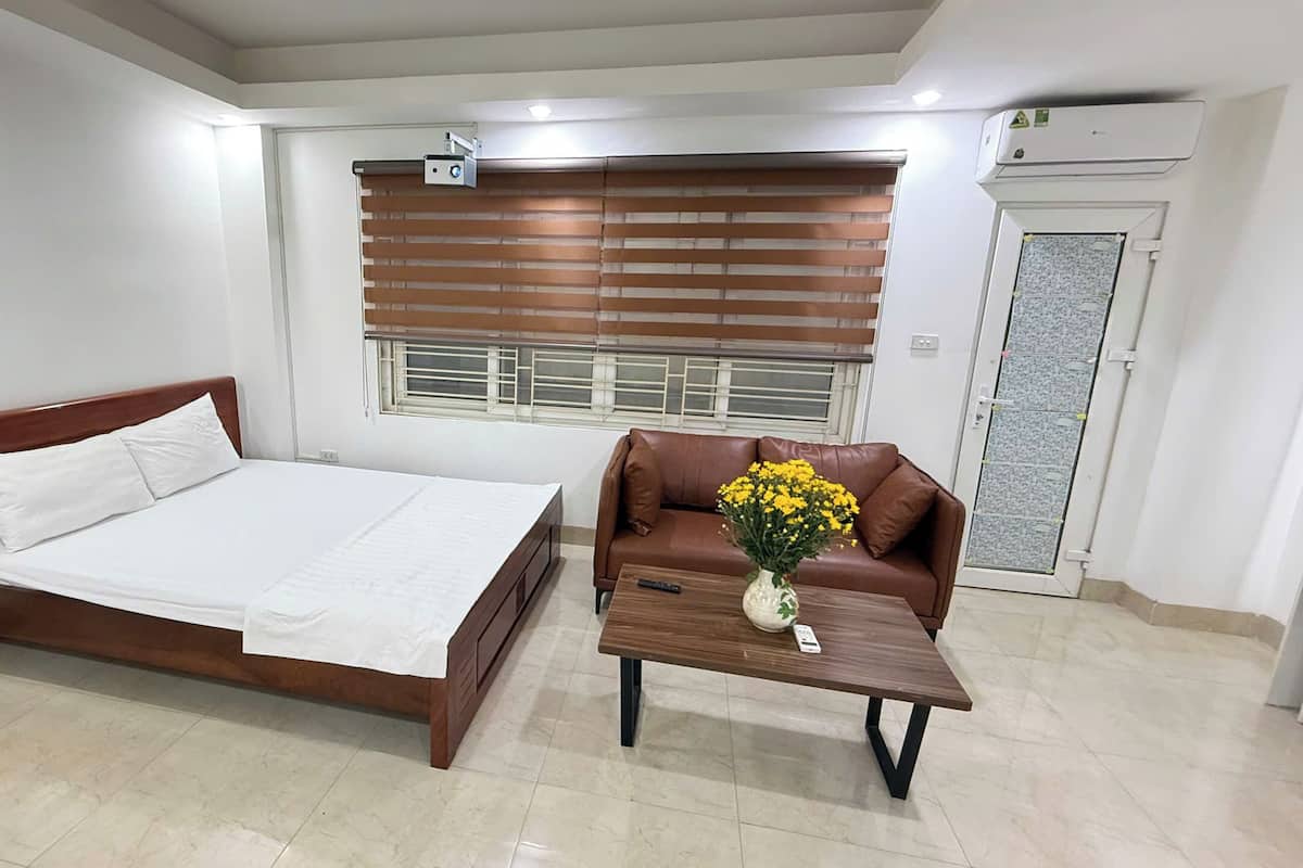 A bedroom with a bed, a brown leather sofa, a small table with a vase of flowers, and a window with blinds.