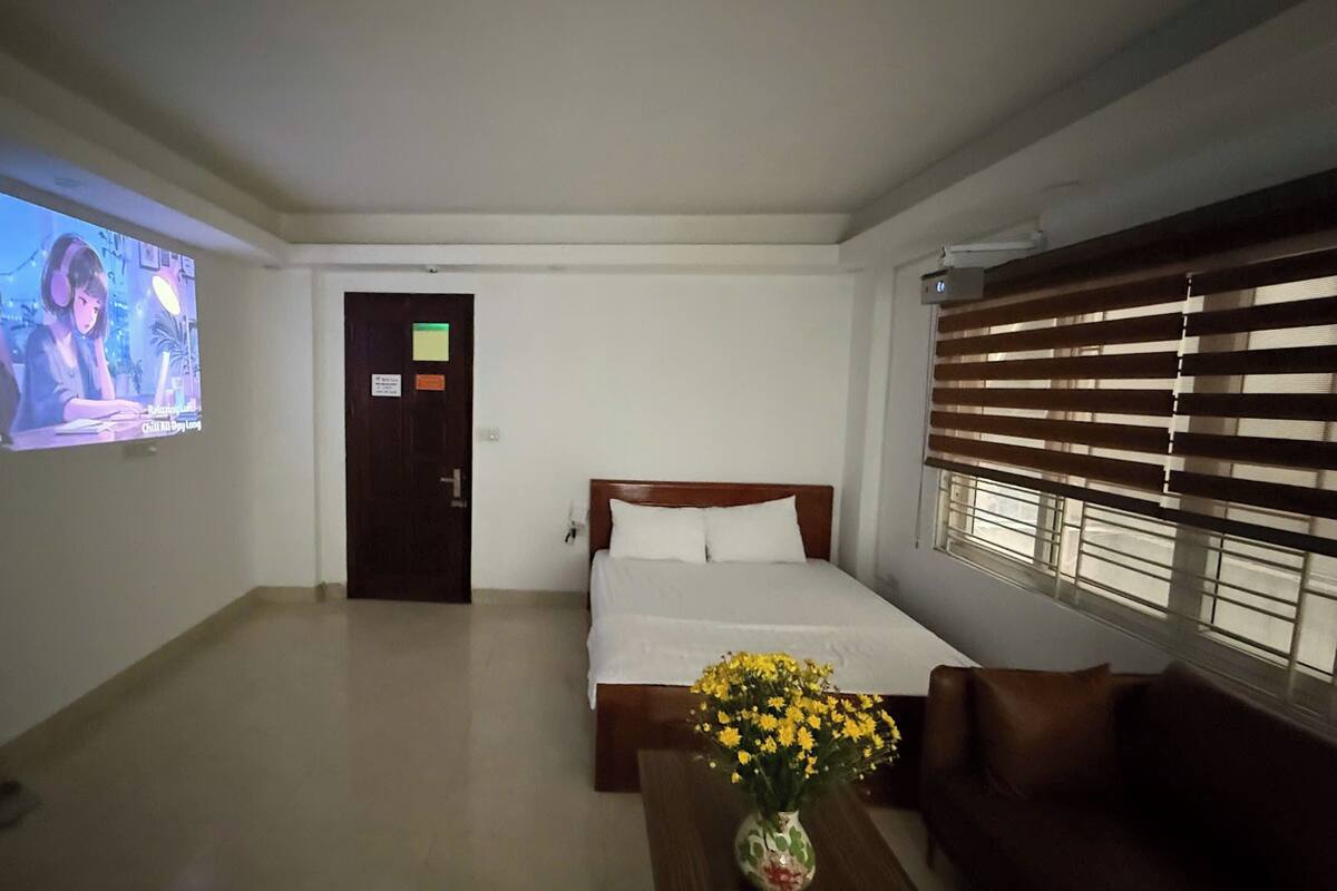 A hotel room with a bed, a small table with a vase of flowers, a television on the wall, and a window with blinds.