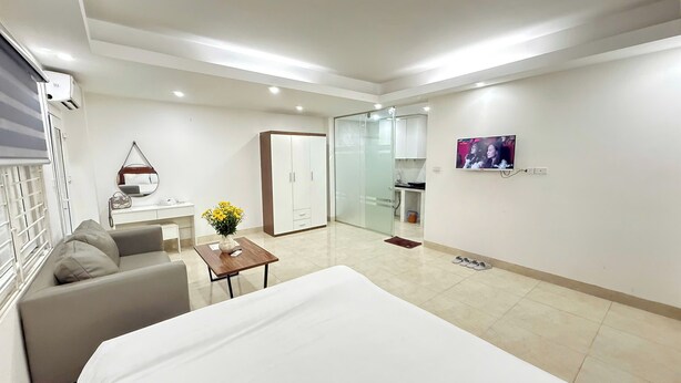 Una habitación de hotel moderna con una cama, un sofá, una pequeña mesa con un jarrón de flores, un armario, una puerta de vidrio que da a un baño y un televisor montado en la pared.