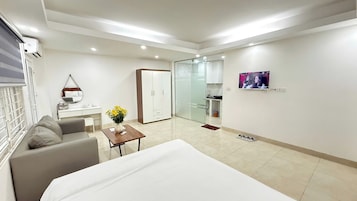 Quarto de hotel moderno com uma cama, um sofá, uma pequena mesa com um vaso de flores, um guarda-roupa, uma porta de vidro para um banheiro e uma TV na parede.