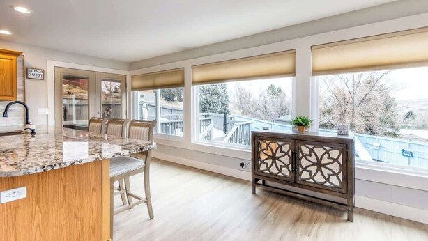 Una cocina con encimera de mármol, armarios de madera, una zona de comedor con sillas y ventanas grandes con persianas.