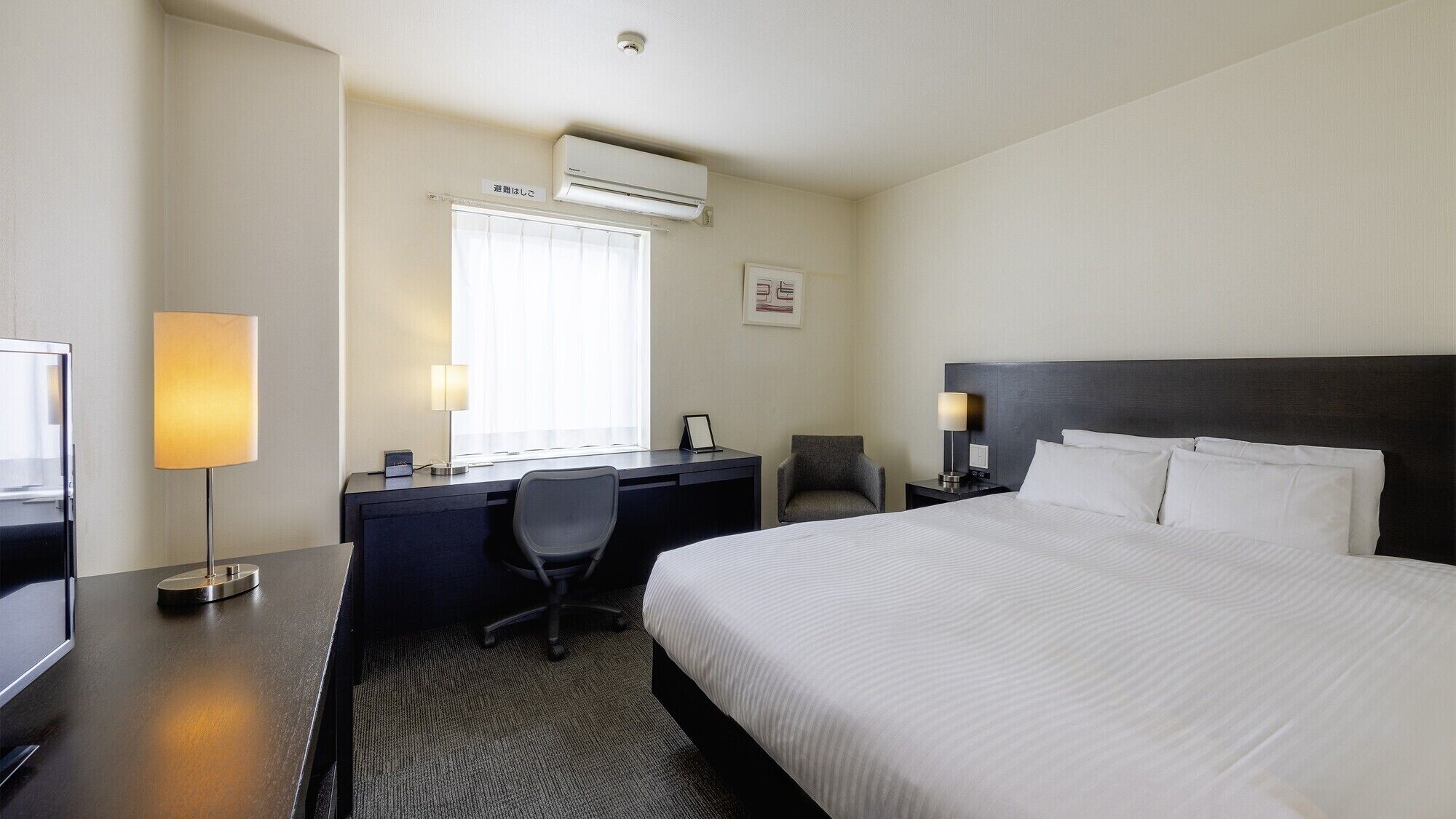 Quarto de hotel com cama, escrivaninha, cadeira e janela com cortinas.